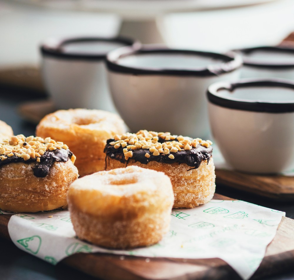 Doughnuts topped with chocolate and crushed peanuts on a serving tray next to coffee cups. 